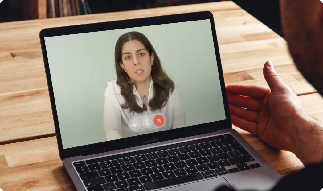 Imagem de uma videochamada sendo exibida em um laptop MacBook Pro. Na tela, uma mulher com cabelos castanhos longos, vestindo uma blusa branca, está falando diretamente para a câmera. A mão de um homem é vista ao lado do laptop, sugerindo que ele está interagindo com a pessoa na videochamada. O laptop está sobre uma mesa de madeira em um ambiente interno.