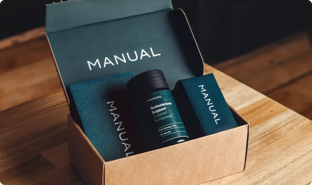 Imagem de uma caixa de papelão aberta sobre uma mesa de madeira, contendo produtos da marca Manual. Dentro da caixa, há um frasco de suplemento com o rótulo ‘Testosterone Support’ e duas caixas menores, também com a marca Manual, todos dispostos de forma organizada.