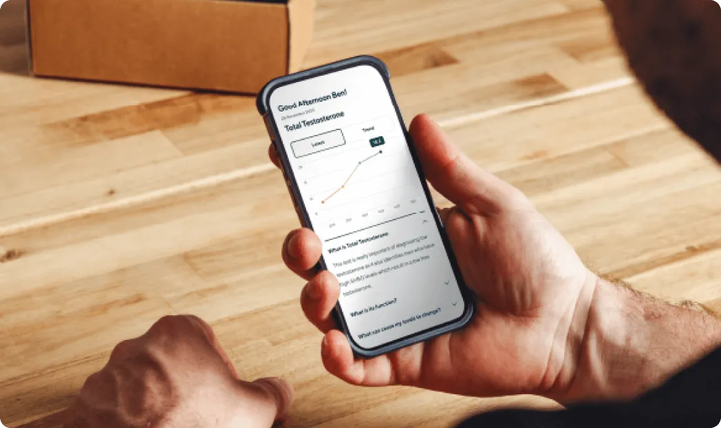 Imagem de uma pessoa segurando um smartphone com a tela exibindo um gráfico de aumento de testosterona ao longo do tempo. Ao fundo, sobre uma mesa de madeira, há uma caixa de papelão fechada.