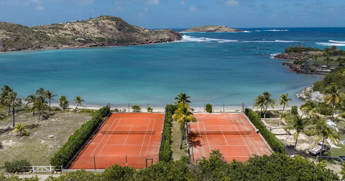 Two clay tennis courts lined with hedges and palm trees, facing a turquoise bay in St Barts