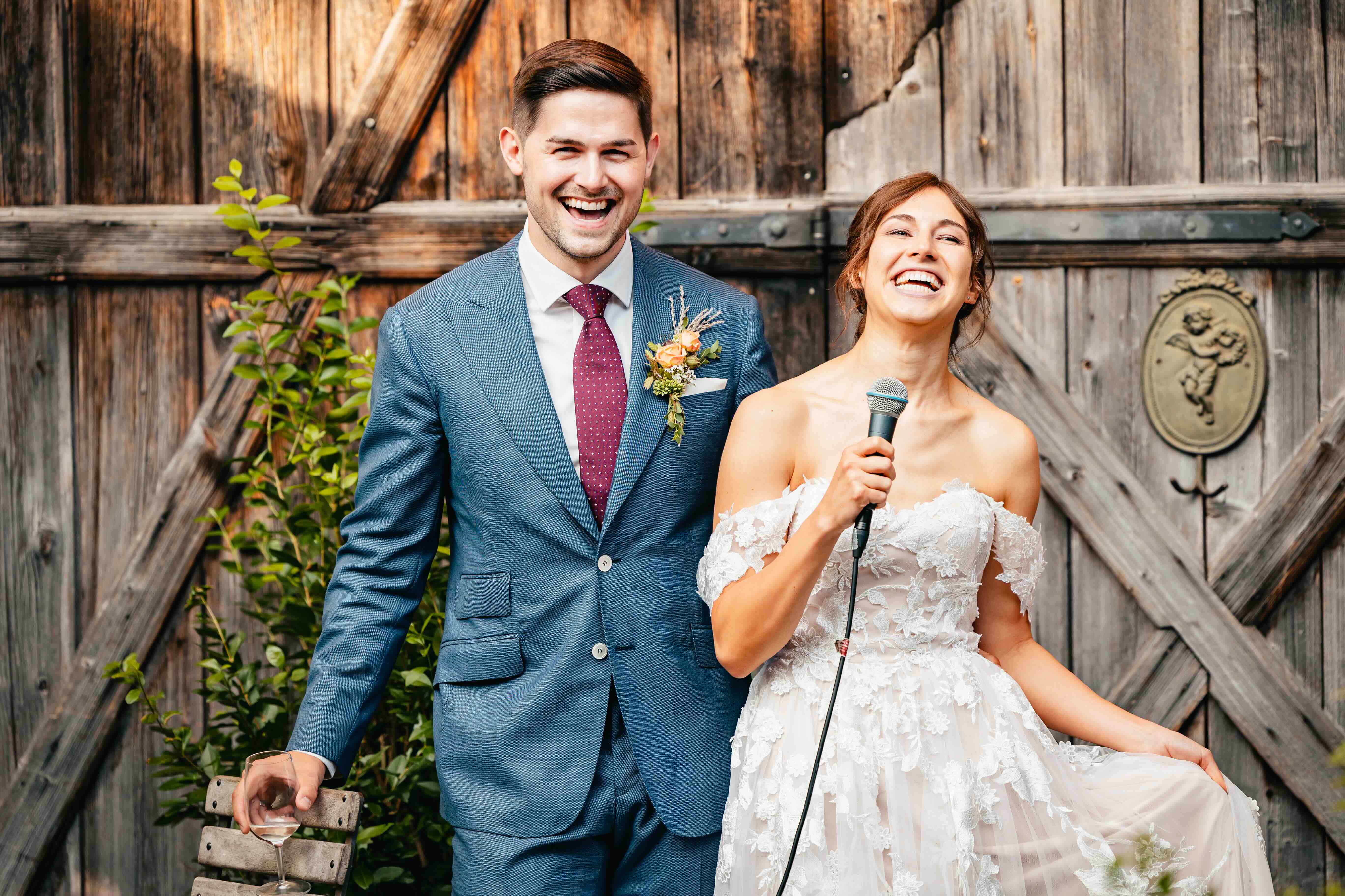 bride and groom laughing while holding microphone in front of wooden fence