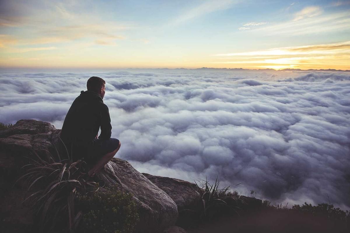 Person sits on rock overlooking clouds.