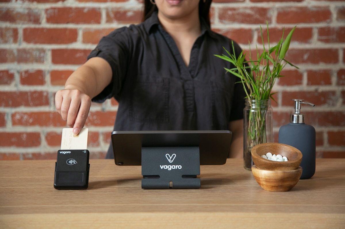 Femme utilisant un terminal de paiement Vagaro sur un comptoir avec des accessoires.