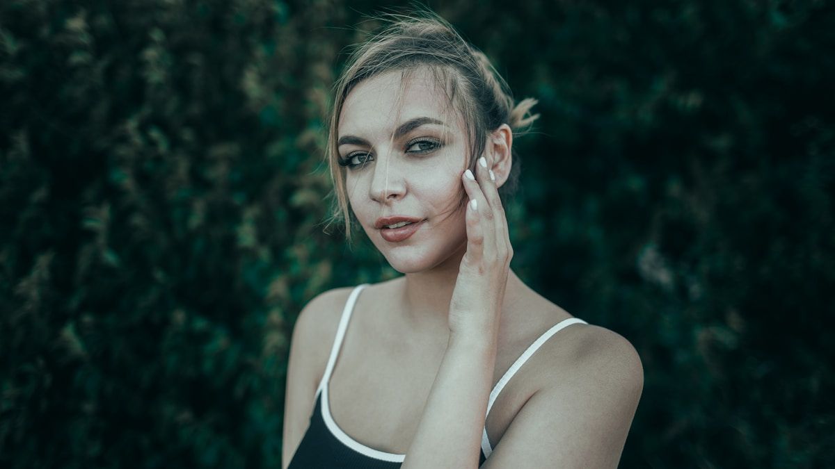 Femme blonde aux yeux perçants, expression pensive dans un environnement forestier.