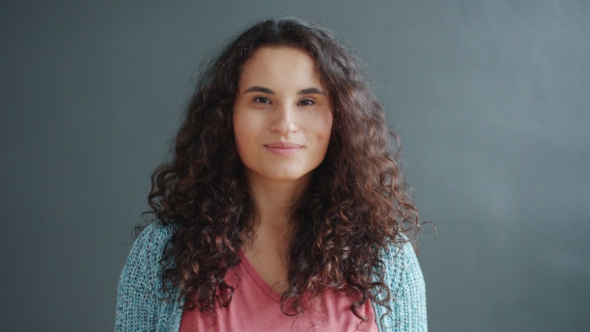 Femme aux cheveux bouclés bruns, souriante, vêtue d'un pull bleu et d'un haut rose.