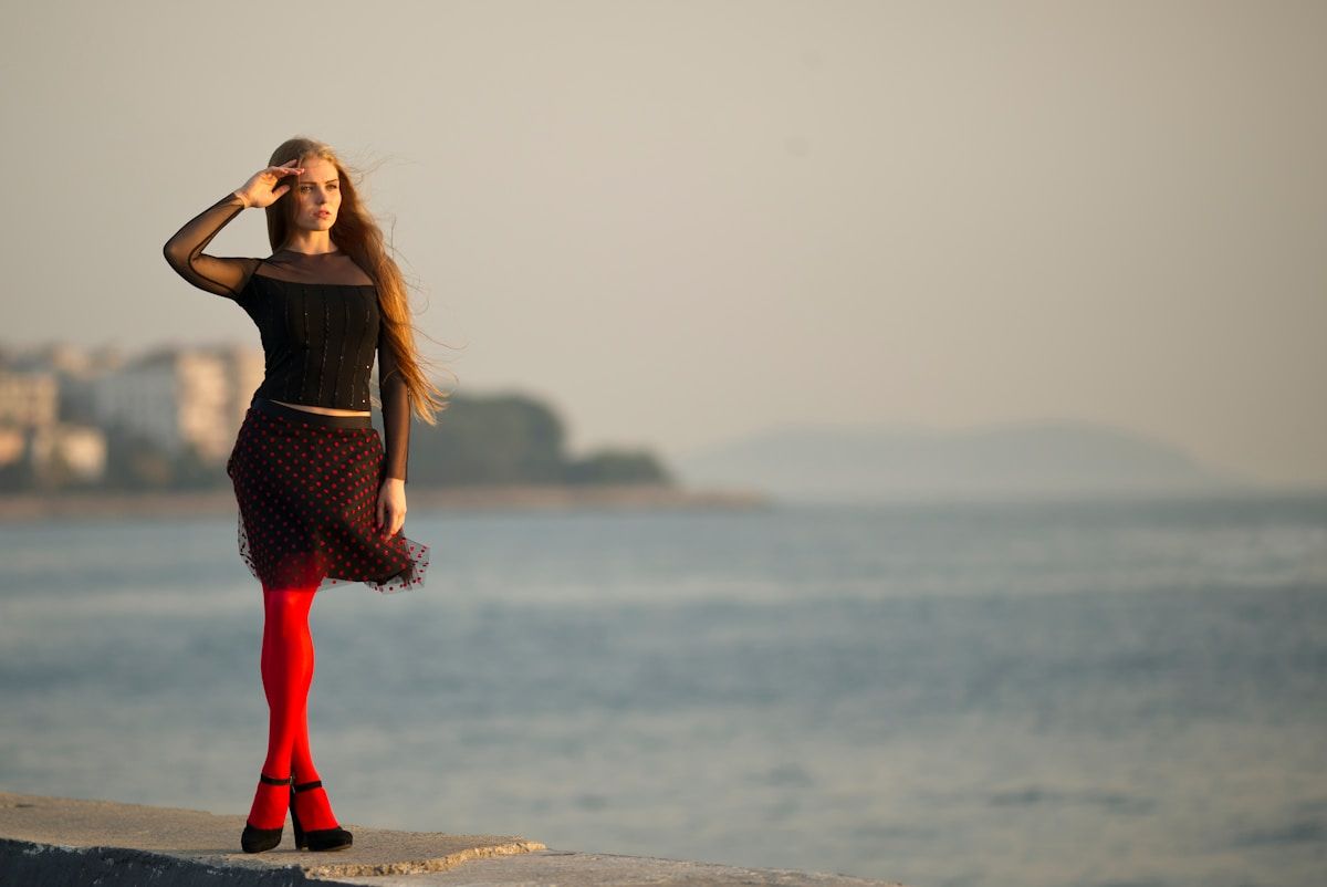 Femme aux cheveux longs debout sur la plage, vêtue d'une jupe à pois et de collants rouges.