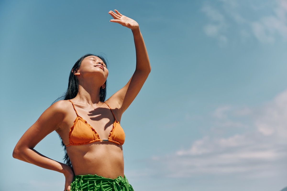 Femme en bikini orange levant les bras sous un ciel bleu.