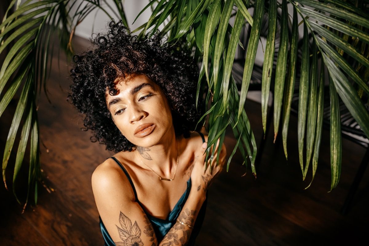 Femme aux cheveux bouclés, tatouages, posant devant des feuilles de palmier.