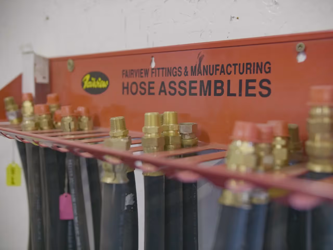 Rack of hydraulic hose assemblies with brass fittings labeled “Fairview Fittings & Manufacturing Hose Assemblies.”