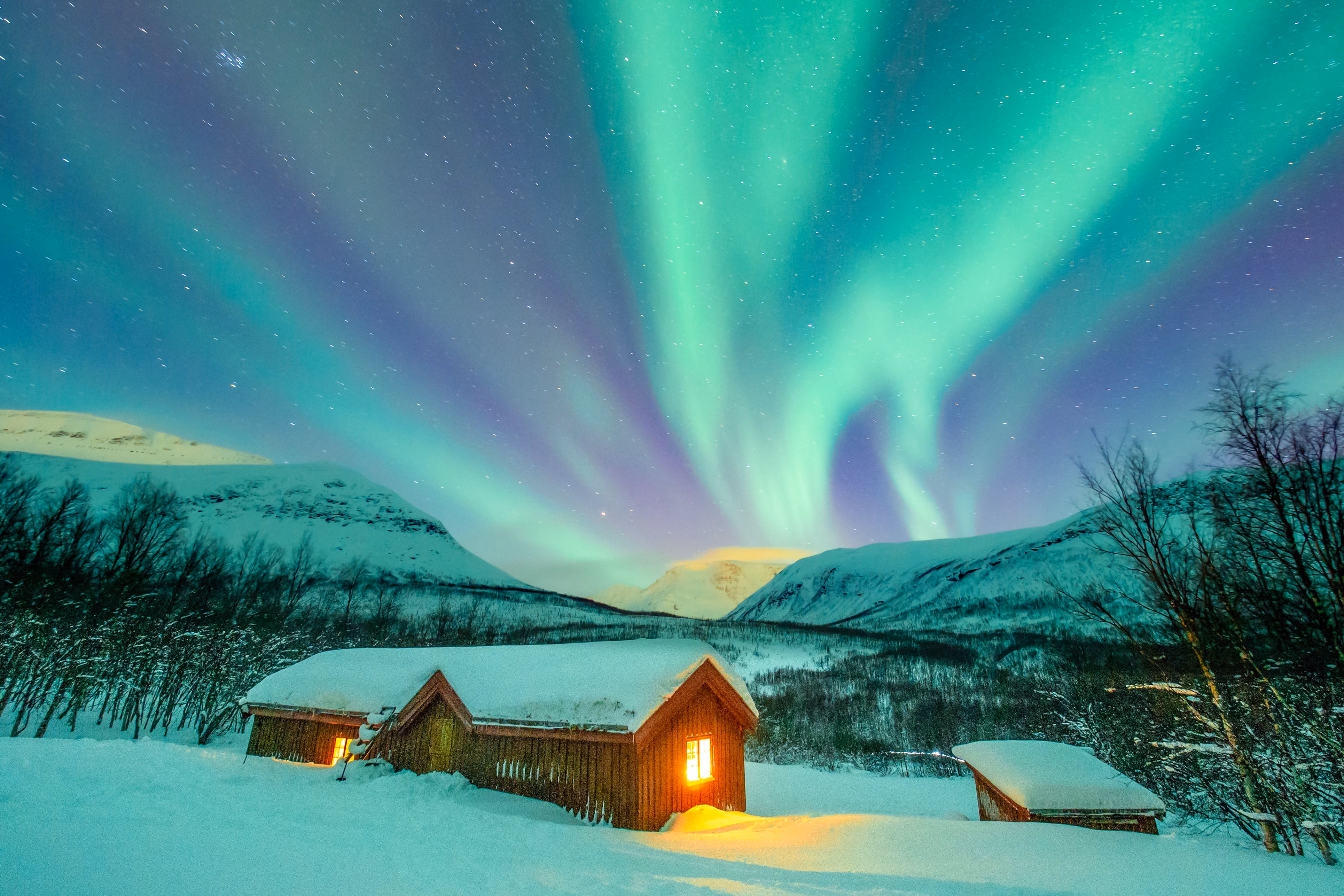 Snow-covered cabins below vibrant green northern lights and starry night sky over snowy mountains.