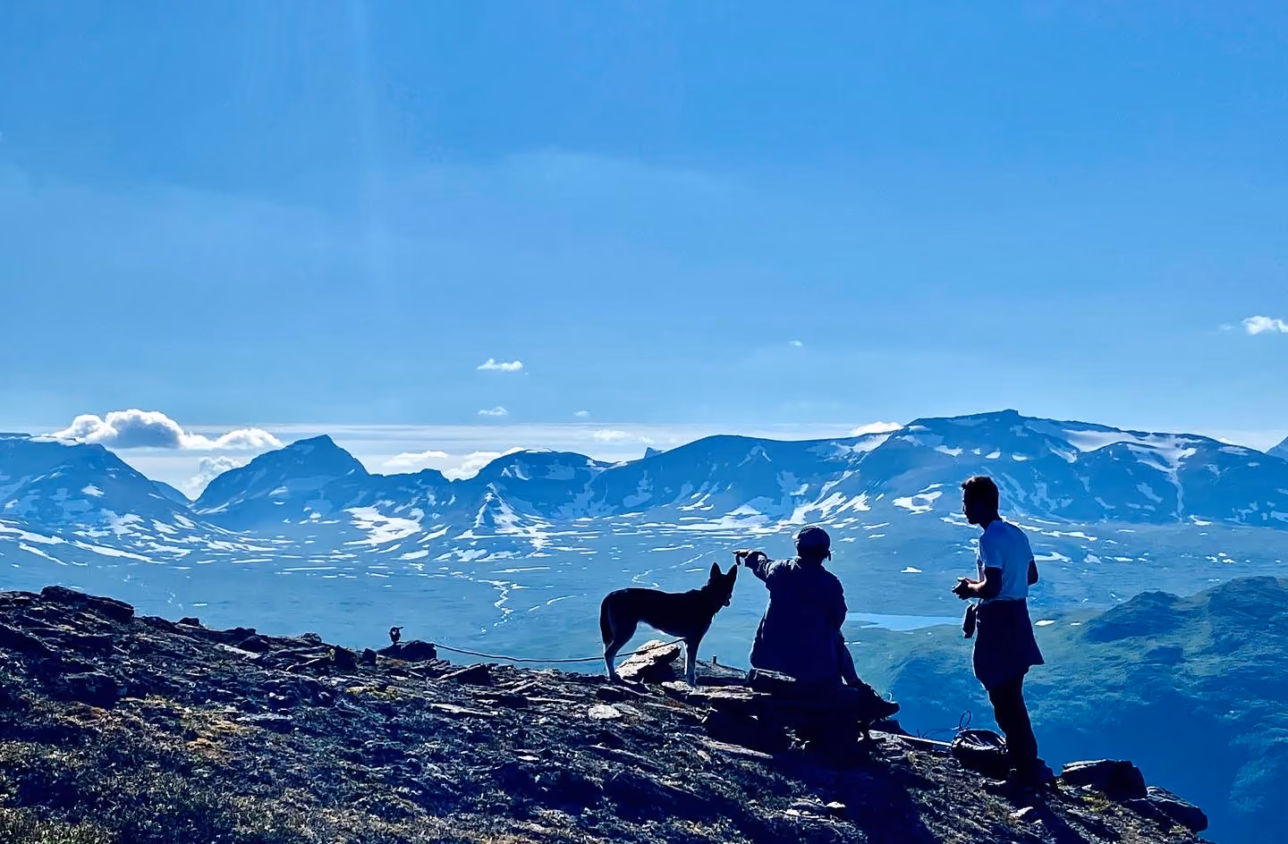 Two people and a dog on rocky terrain with snowy mountain range in the background under a clear sky.