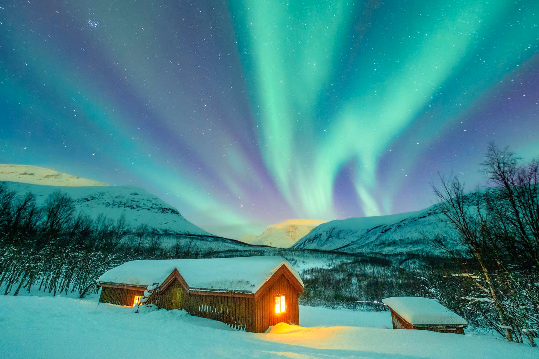 Snow-covered cabin with warm glowing windows under green northern lights and starry sky in mountainous landscape.
