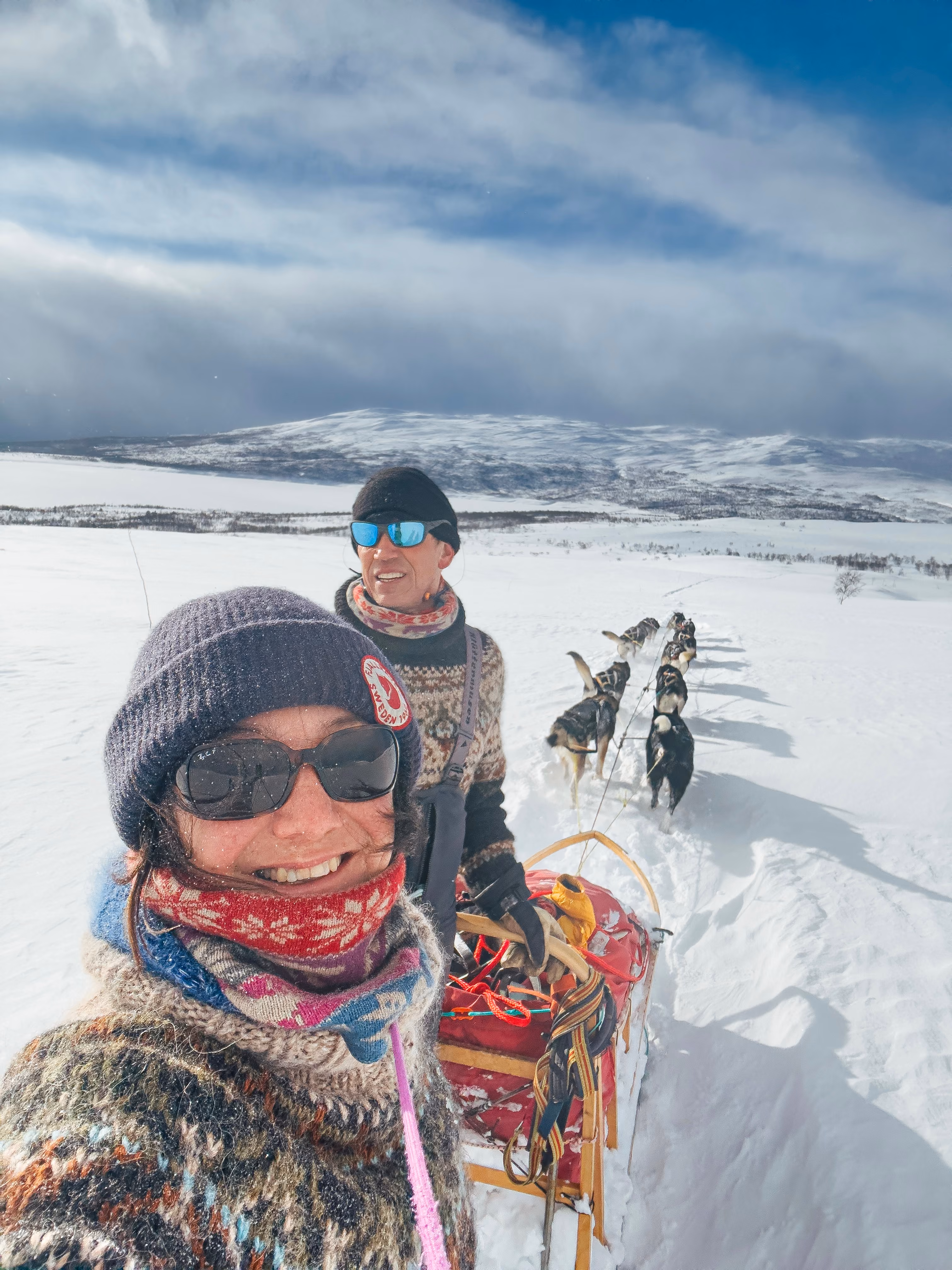 Two people smiling on a dog sled ride with huskies across snowy mountains under cloudy sky.