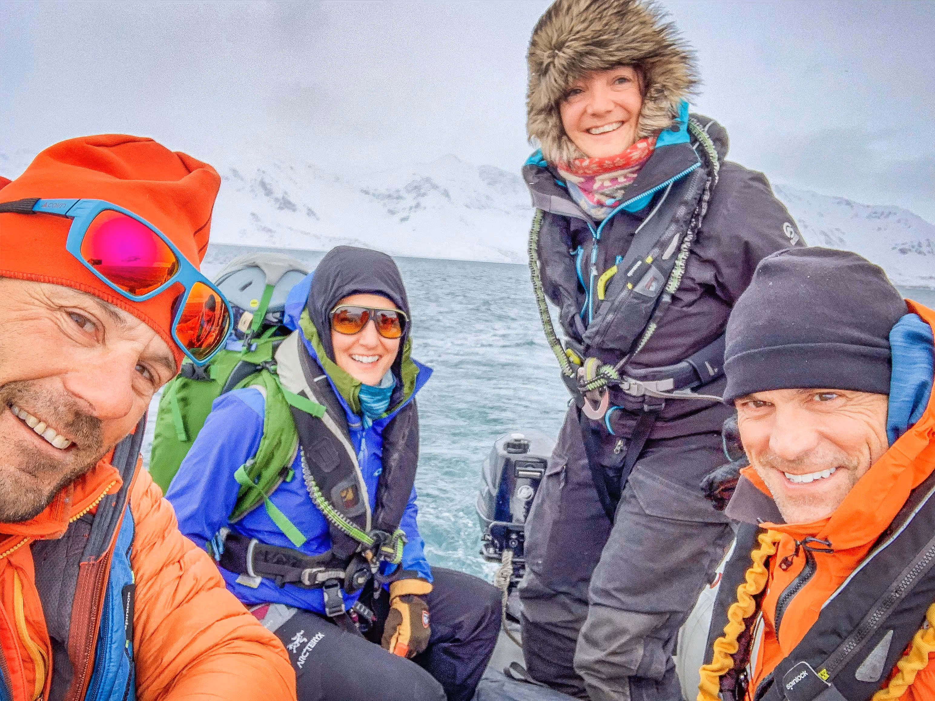 Four people in colorful winter gear on a boat in icy waters with snowy mountains in the background.