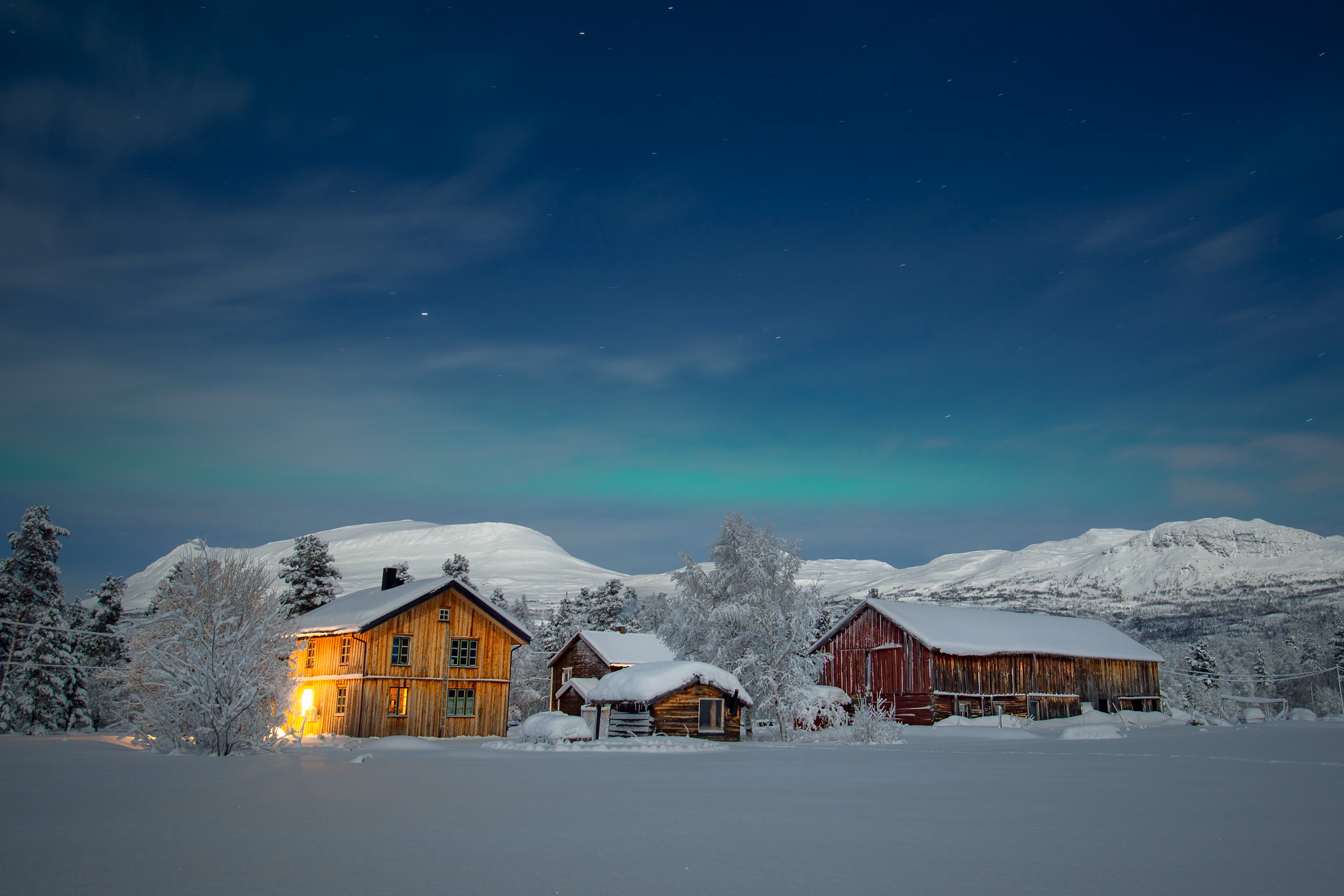 Snow-covered wooden houses lit warmly at night under northern lights in a mountainous winter landscape.