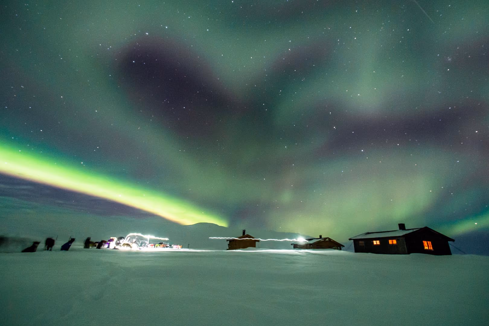 Northern lights glowing green above snowy ground with lit cabins and light trails.