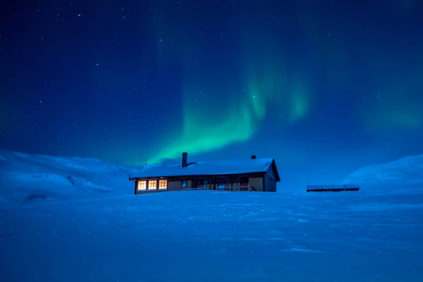Snow-covered cabin at night with lit windows under green northern lights in a starry sky.