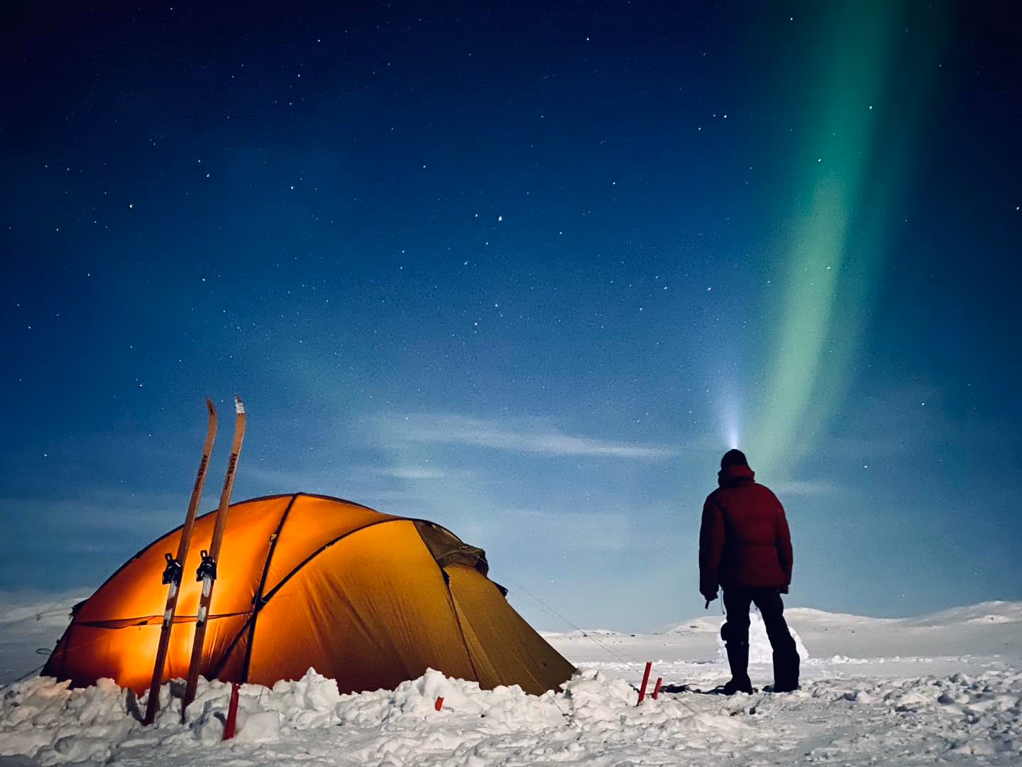Person with headlamp stands in snowy landscape near lit orange tent under starry sky with northern lights.