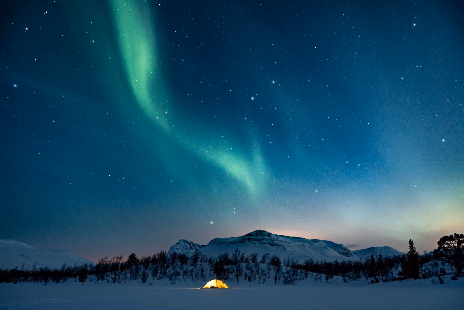 Glowing yellow tent on snowy field beneath green northern lights and star-filled night sky.