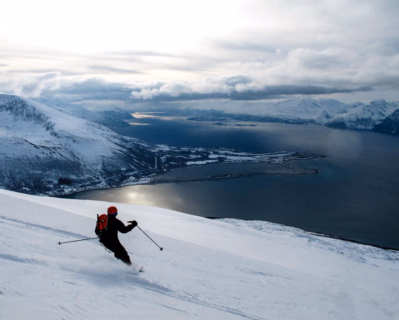 Skier skiing down a snowy slope with a scenic ocean fjord below.