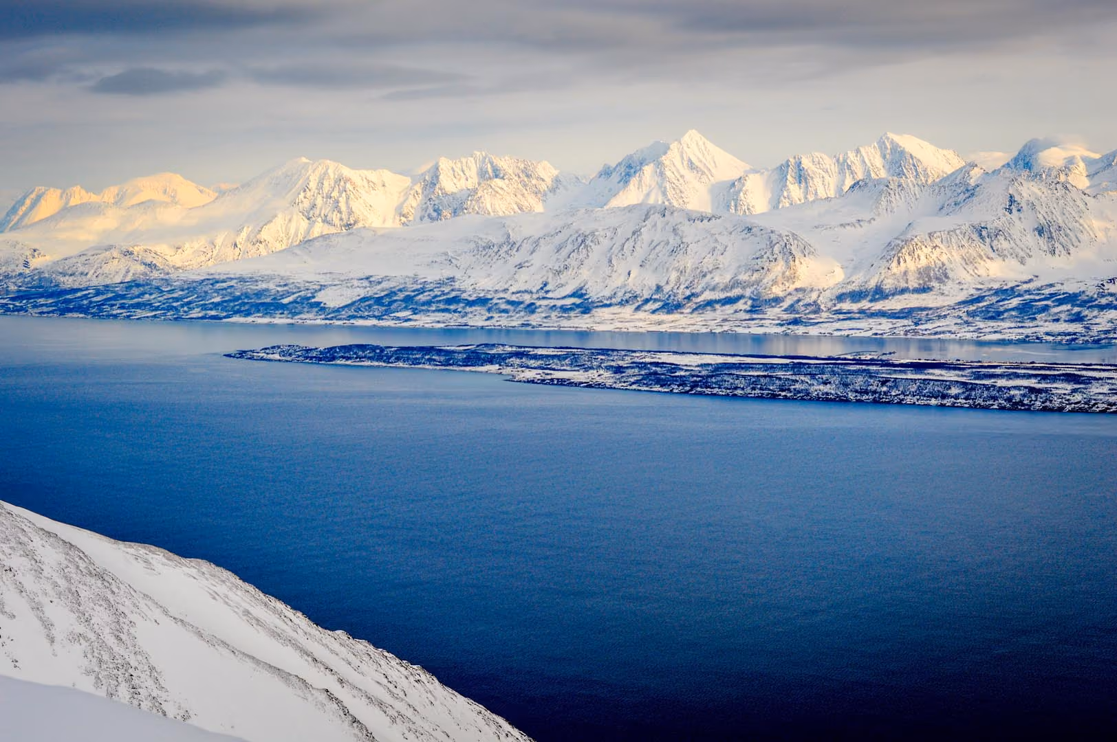 Wide view of a deep blue fjord surrounded by snow-capped mountain ranges.