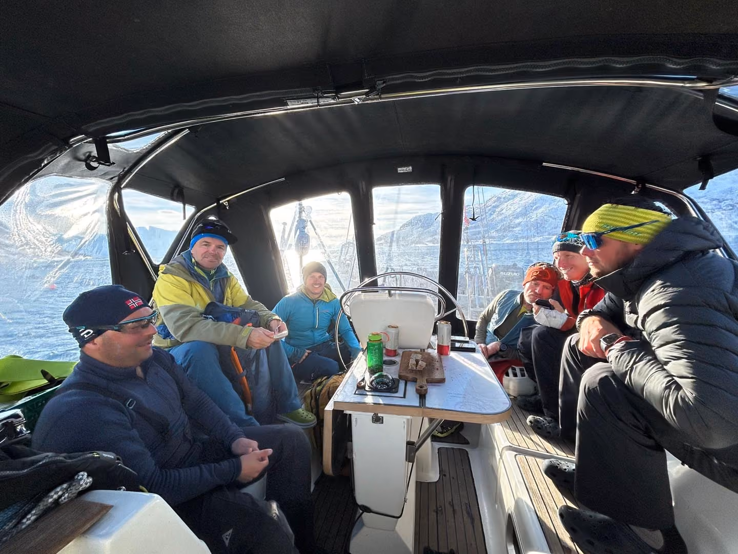Six people dressed warmly seated inside a covered sailboat cockpit on water with mountains outside.