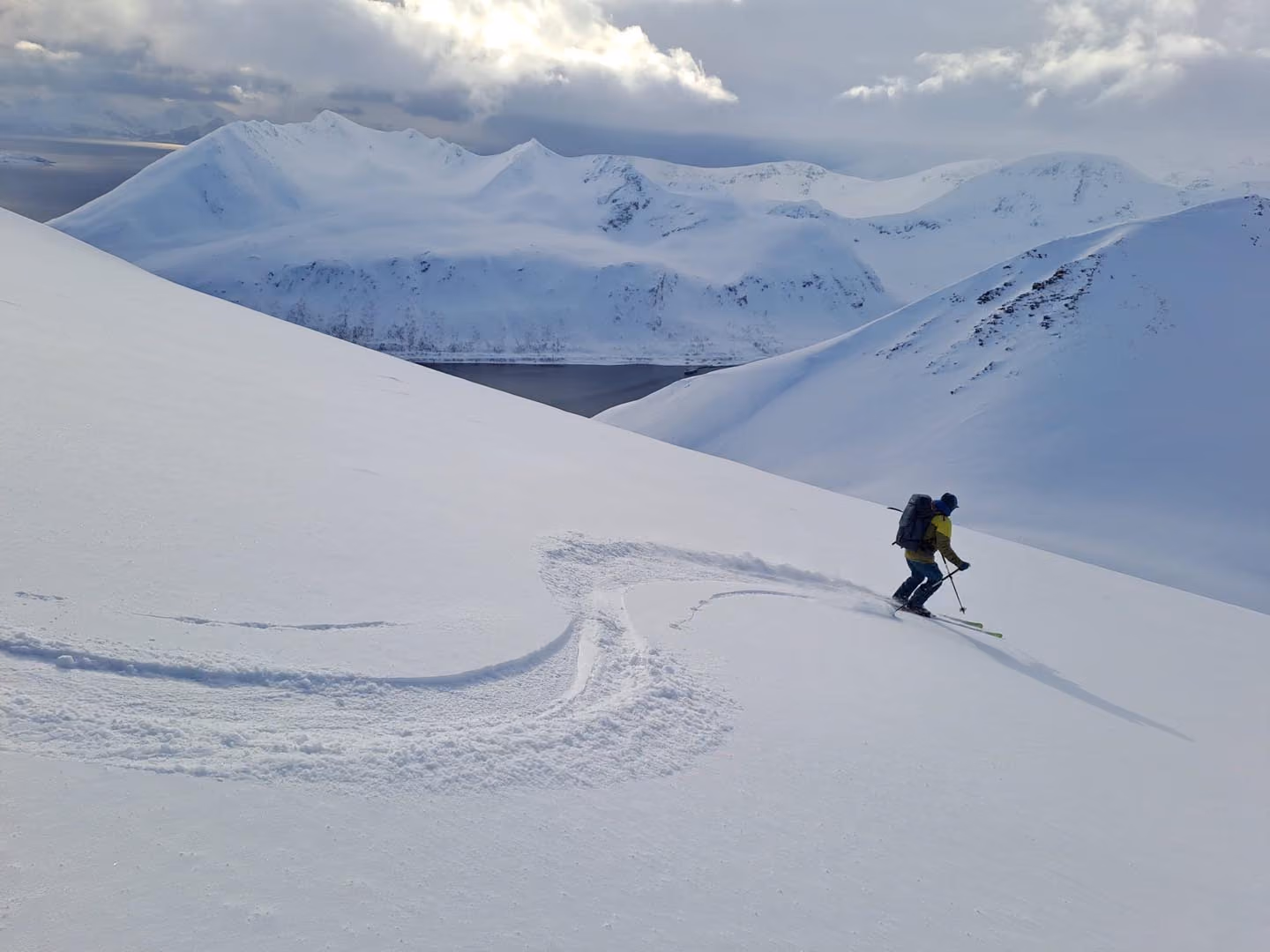 Lone skier carving turns down a steep snowfield toward a fjord.