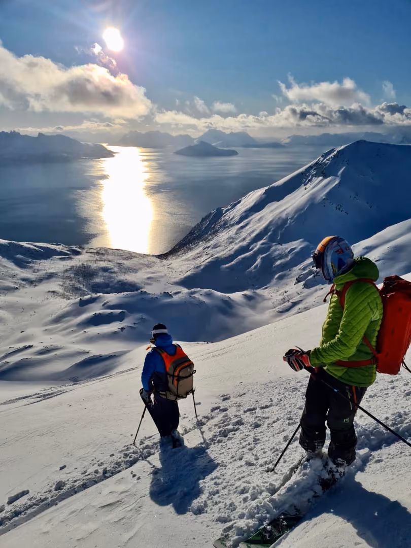 Two skiers descending a snowy mountain with a sunlit sea and islands in the background.