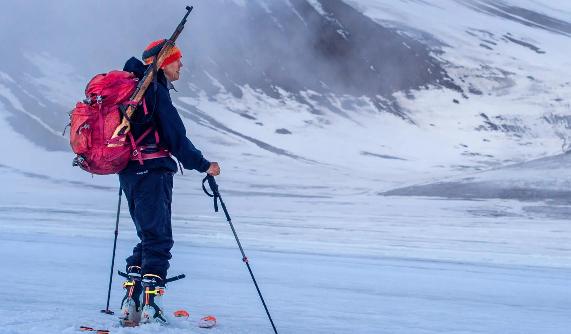 Person skiing on snow-covered mountain with skis, poles, red backpack, and rifle strapped to backpack.