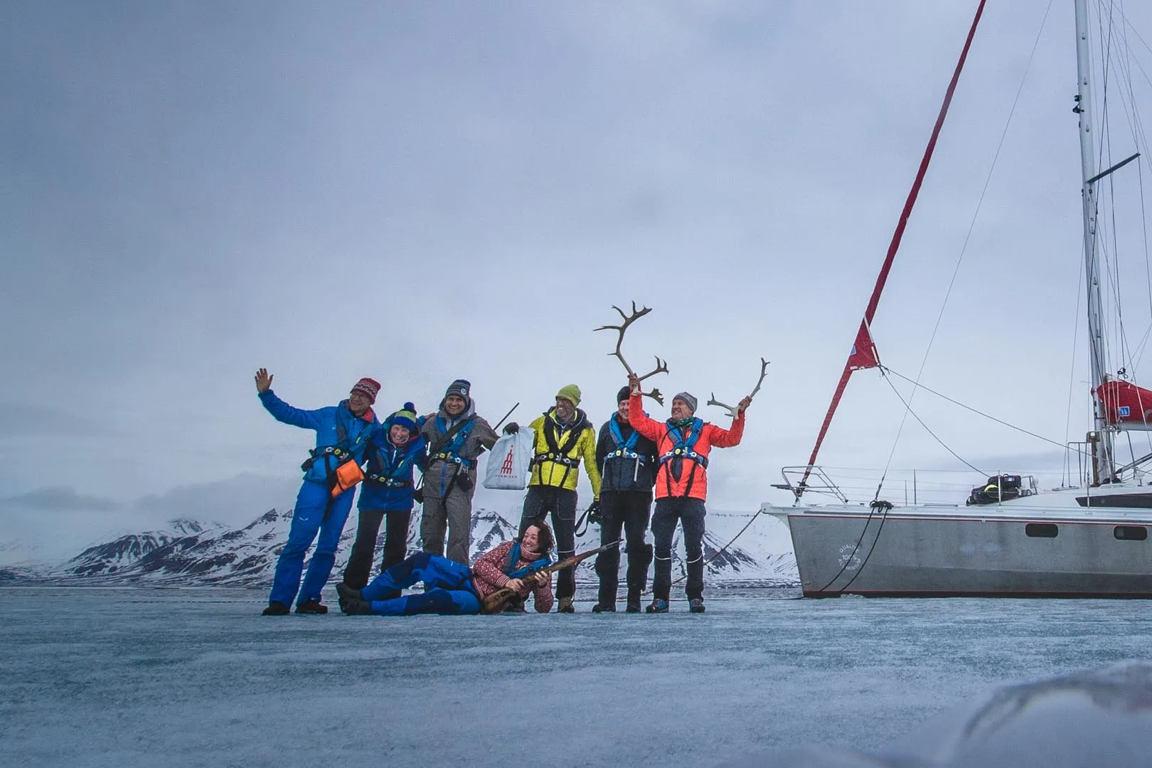 Group of seven people in winter clothes on snow with antlers and a boat nearby.