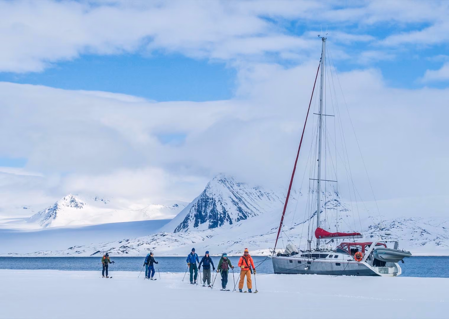 Group skiing on snow near a sailboat with snowy mountains and blue sky in the background.