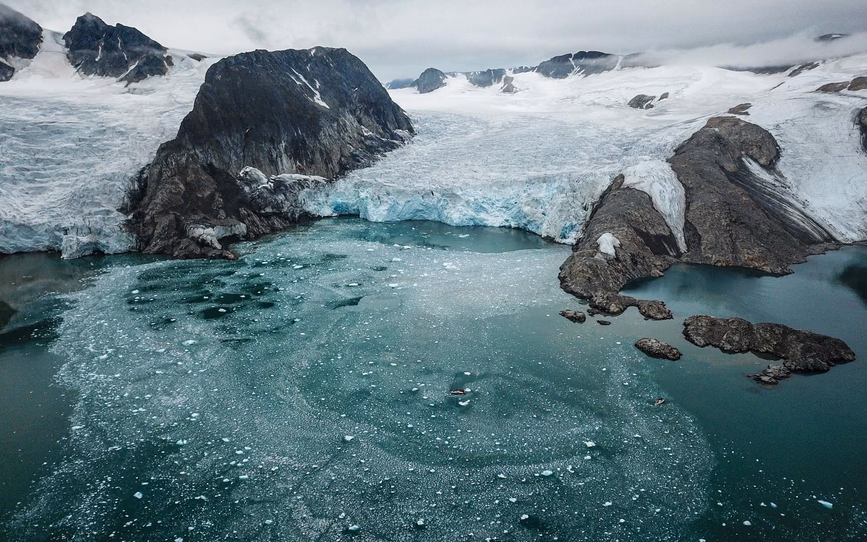 Icy fjord with melting glacier, floating ice chunks, and rocky mountain slopes under cloudy sky.