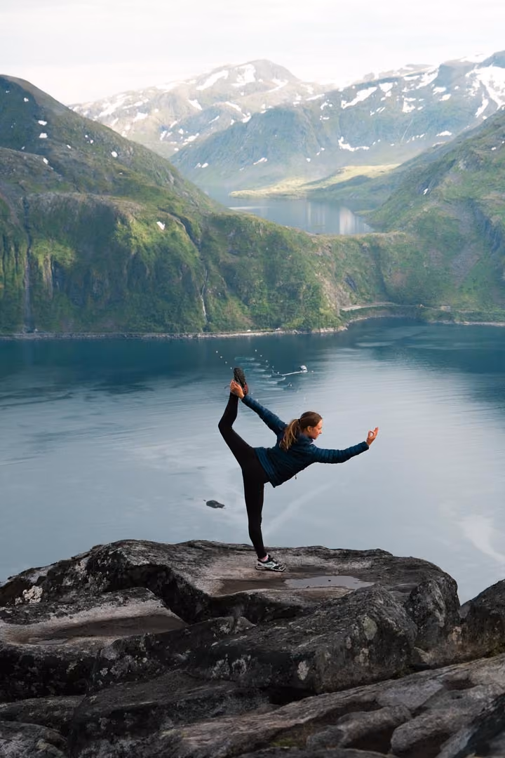 Woman in jacket and leggings balancing on one leg on rocky cliff above lake and mountains.