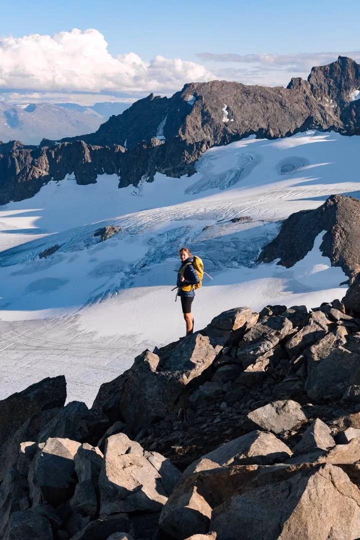 Person hiking along a rugged mountain trail with a tarn nestled between peaks.