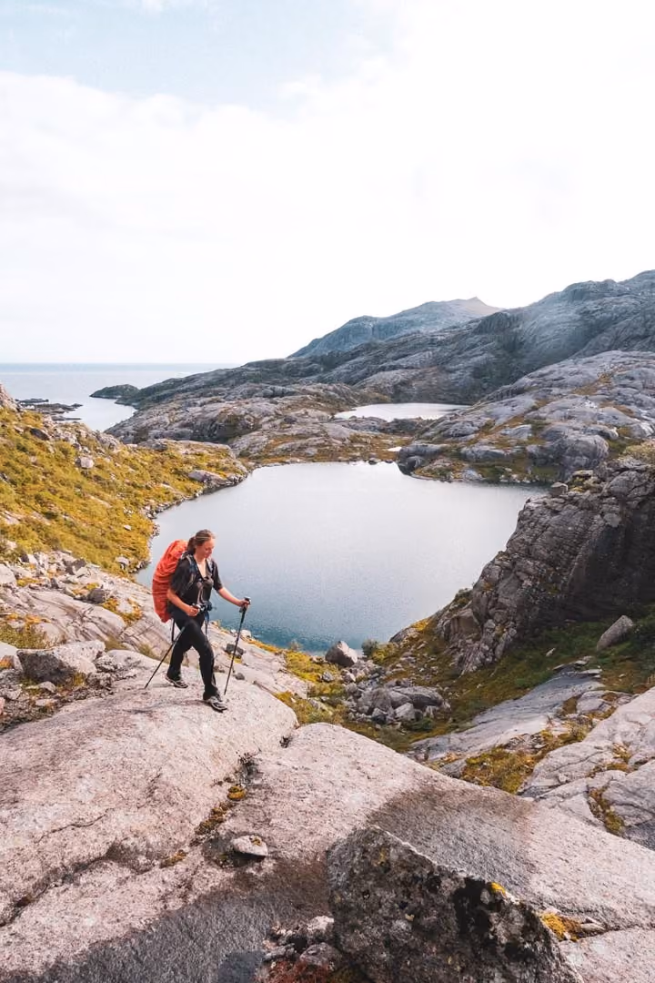 Person hiking along a rugged mountain trail with a tarn nestled between peaks.