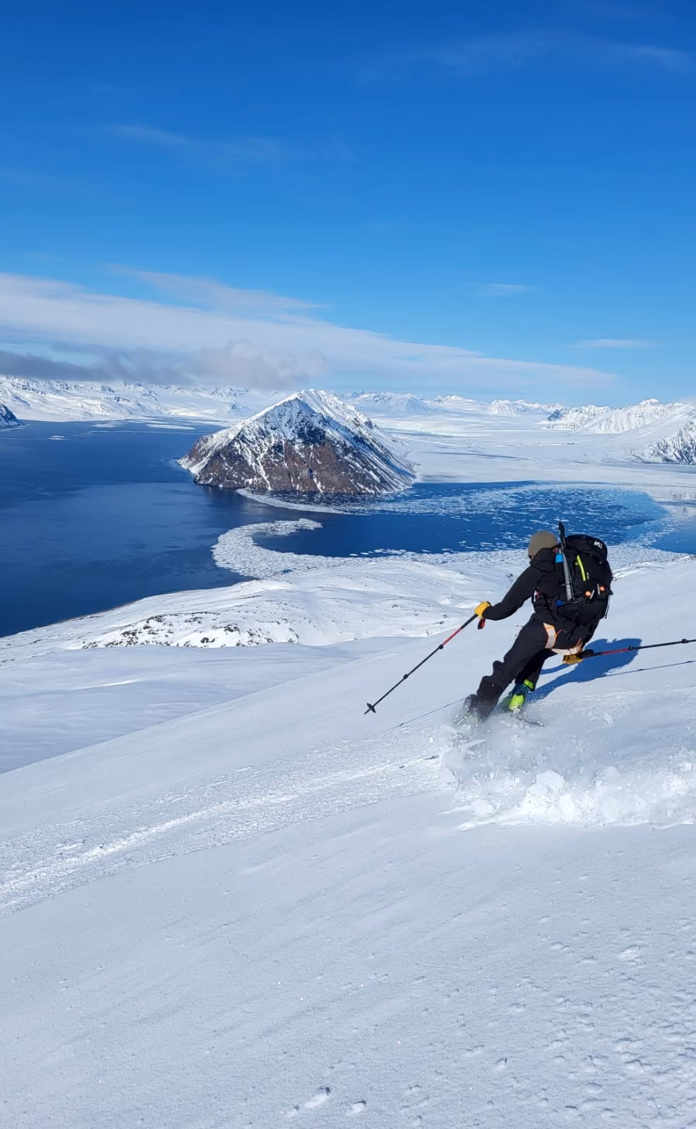 Skier in black gear skiing down a snowy mountain slope with icy water and snow-covered peak behind.