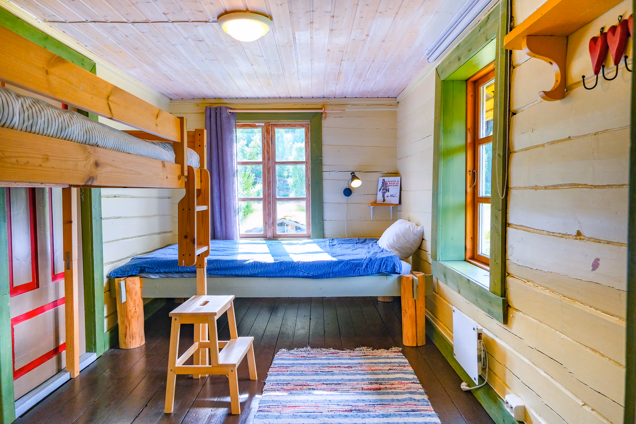 Cozy wooden room with bunk bed, single bed with blue bedding, small stool, and striped rug on floor.