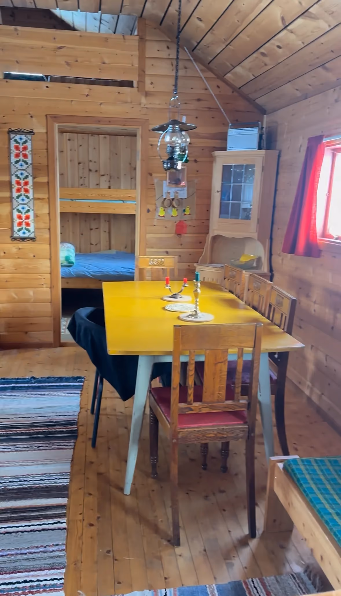 Wood-paneled dining room with a yellow table, wooden chairs, hanging lamp, and bunk bed in the background.
