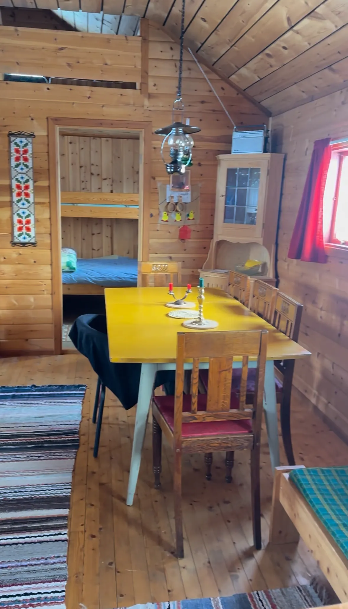 Wood-paneled dining room with a yellow table, wooden chairs, hanging lamp, and bunk bed in the background.