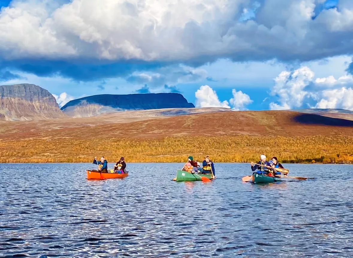 Group of canoes paddling together on a blue lake with autumn hills in the background.