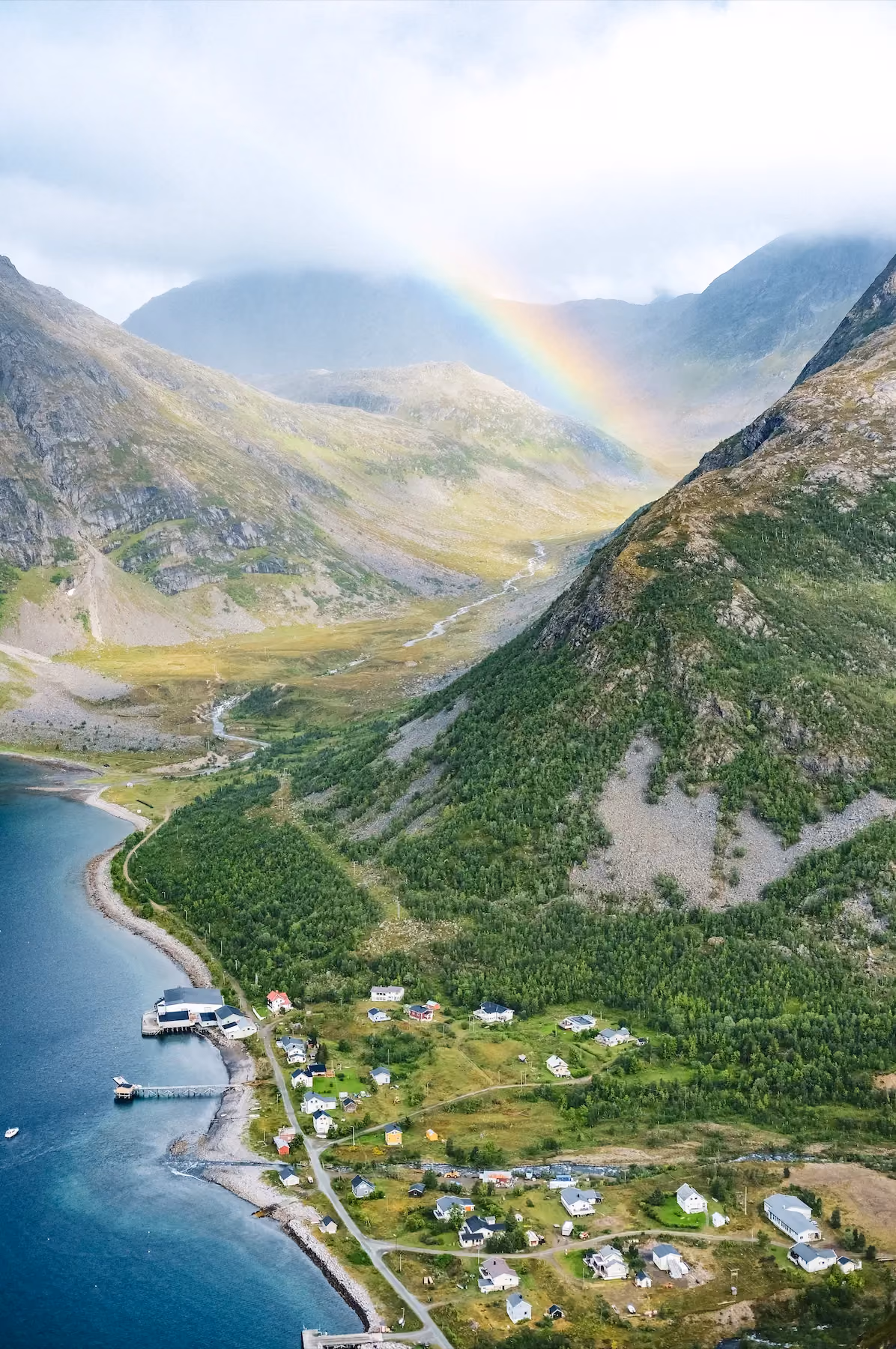 High aerial view of steep mountain ridges and a fjord under a soft rainbow.