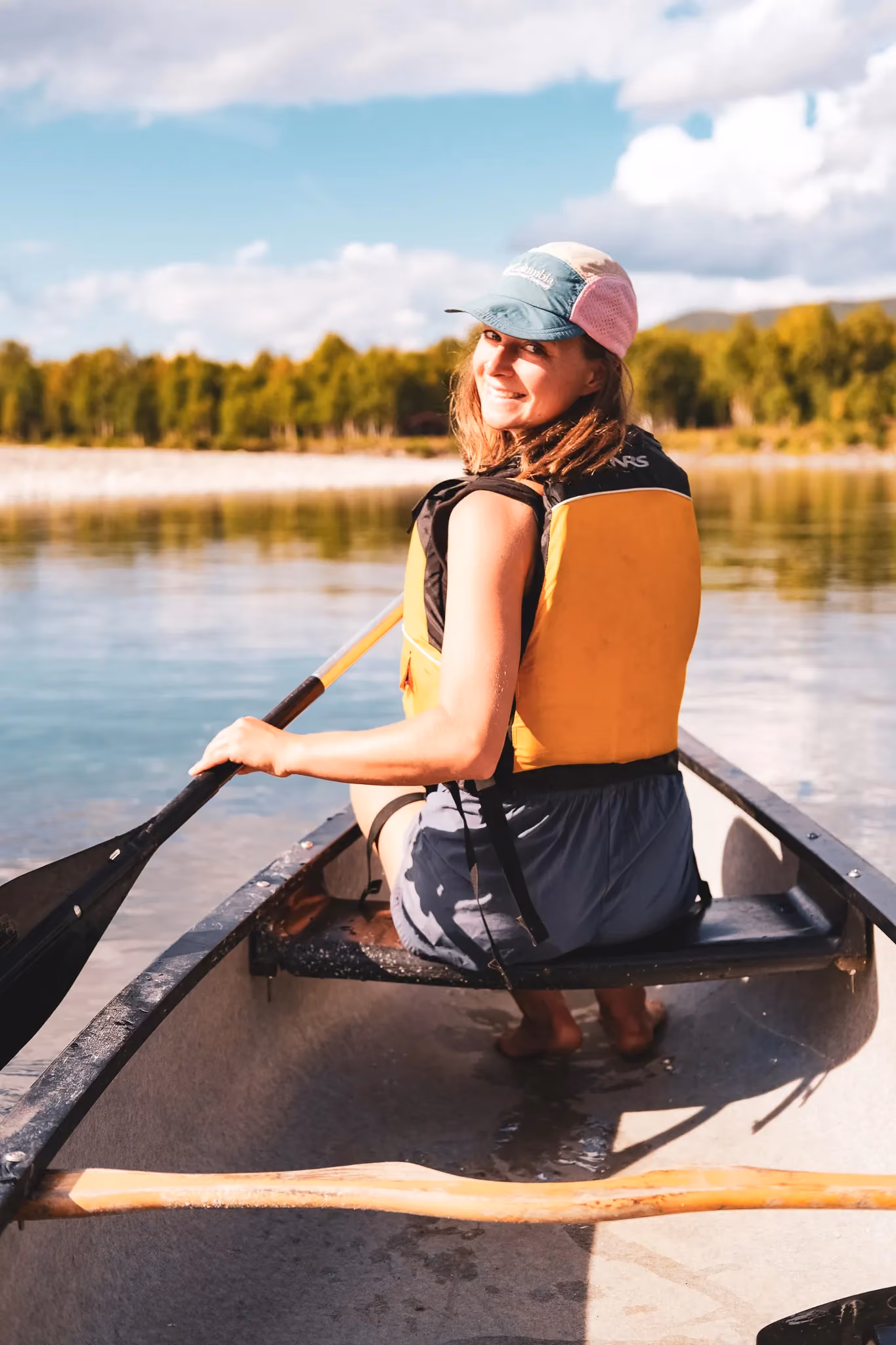Person wearing a life vest paddling a canoe on a calm, sunlit lake.
