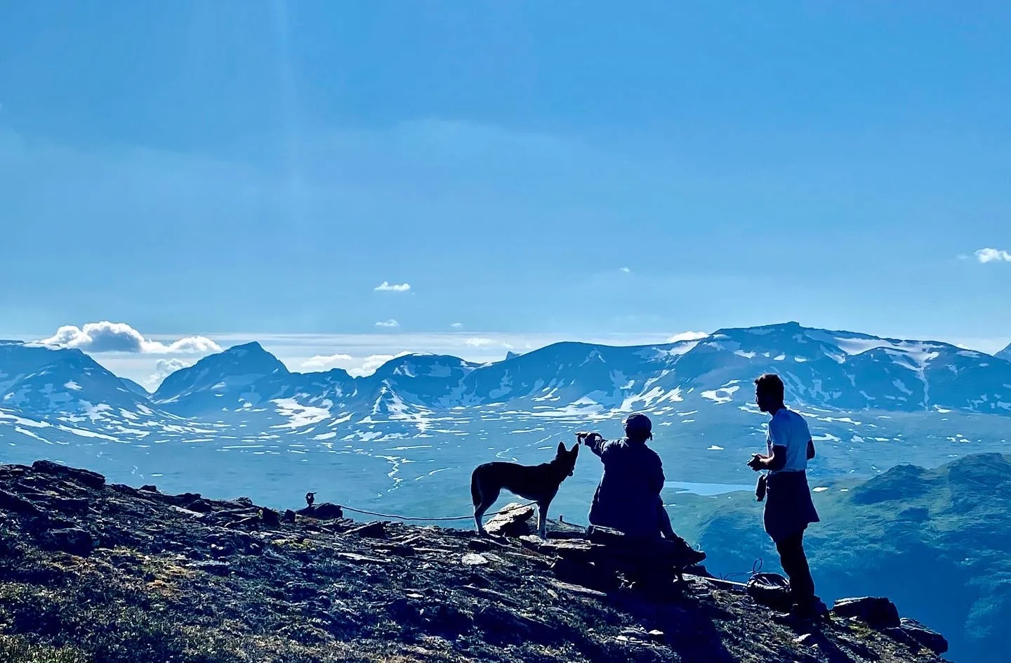 Two people with a dog on rocky terrain overlooking snow-capped mountains under a clear sky.
