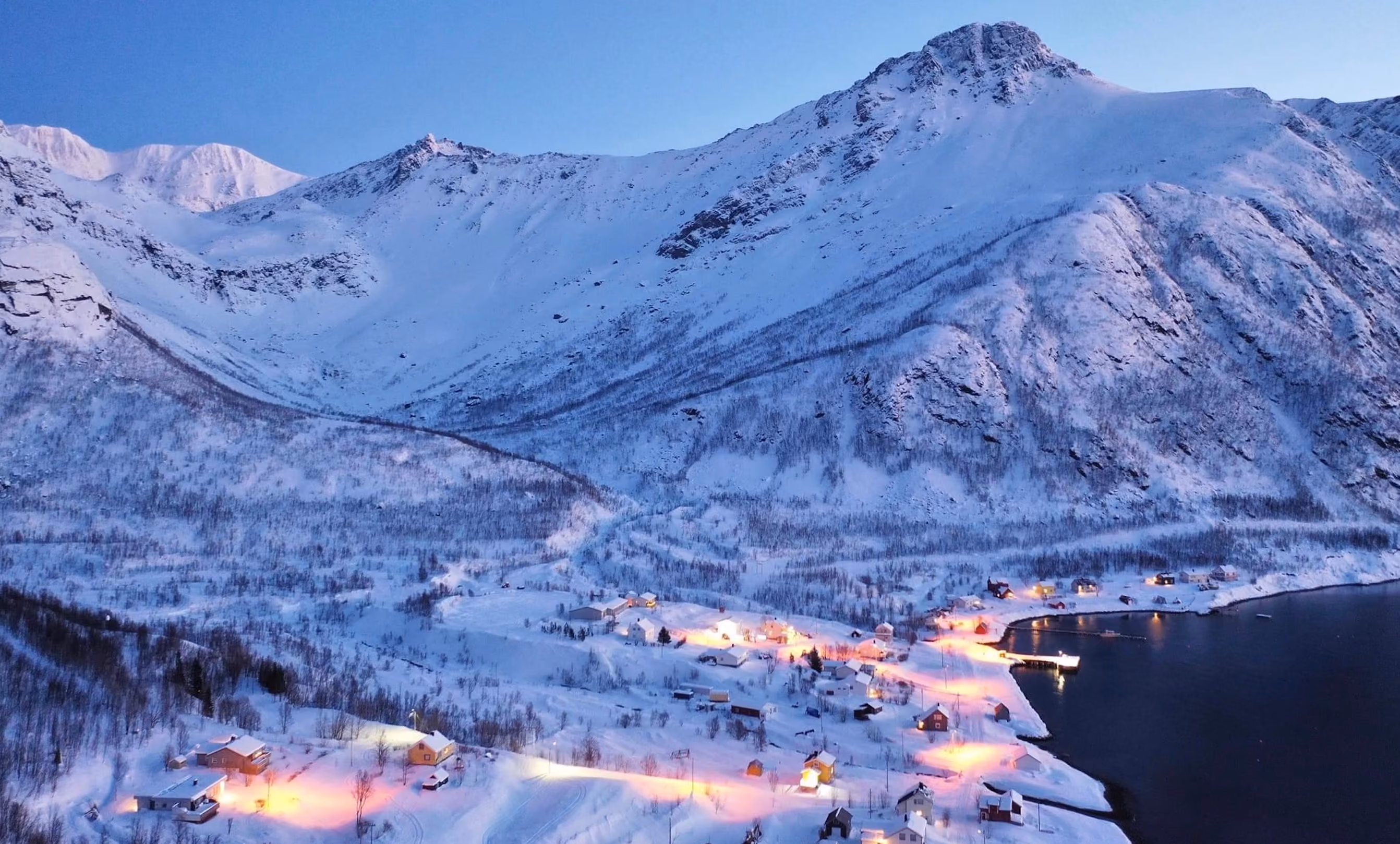 Snow-covered village with lit houses by a fjord and large snowy mountains under a clear blue sky.