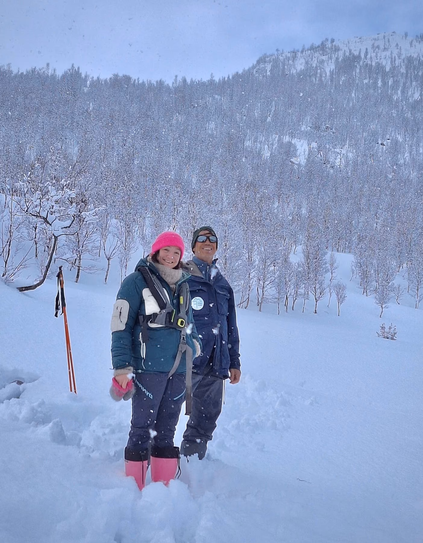 Two people smiling in deep snow with snowy trees and mountain in the background.