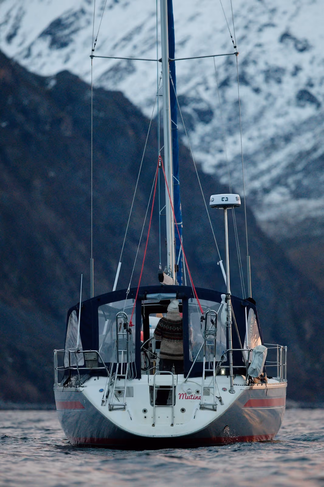 Rear view of a sailboat docked in water beneath steep, dark mountain cliffs.