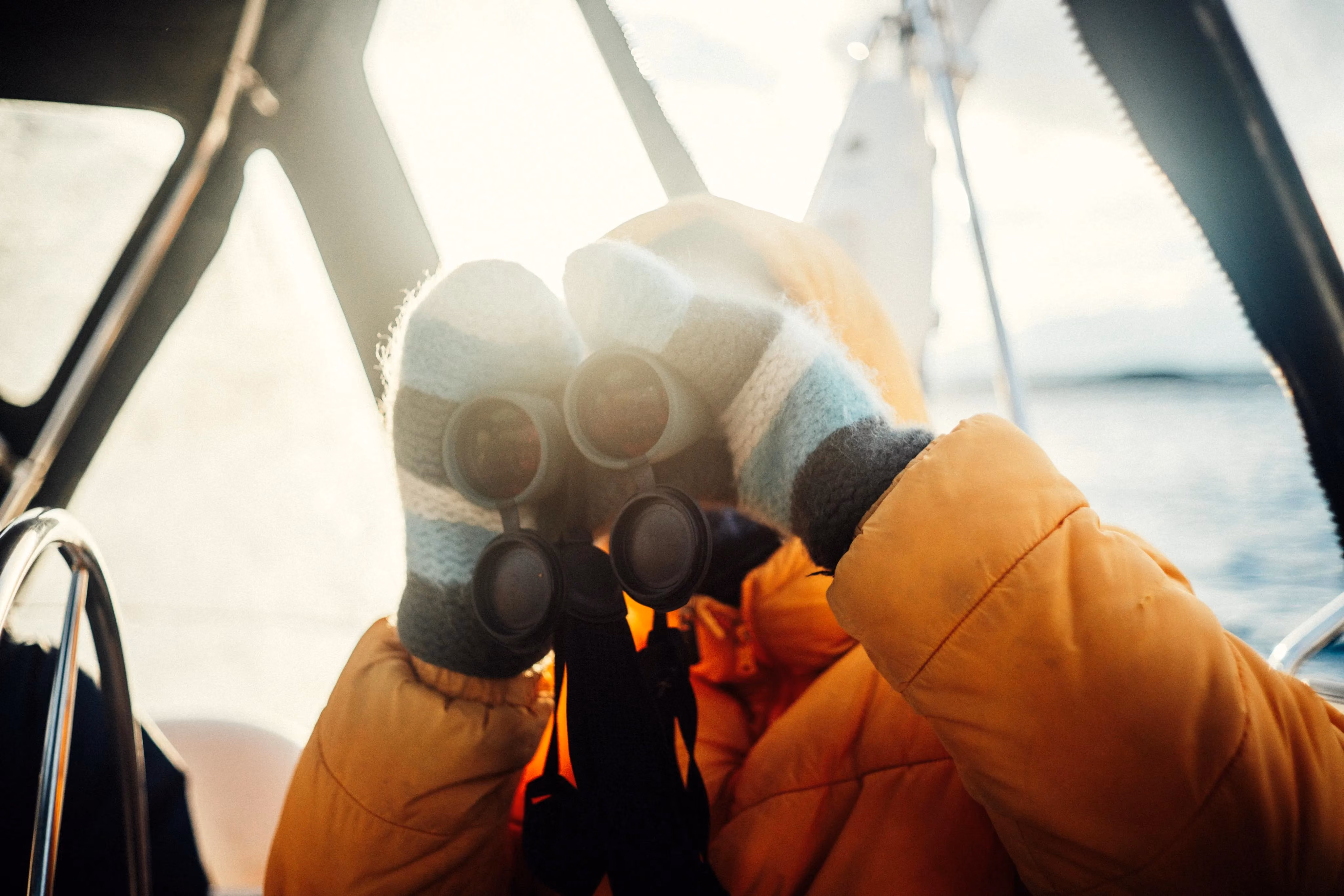 Person in an orange winter jacket looking through binoculars on a boat deck.