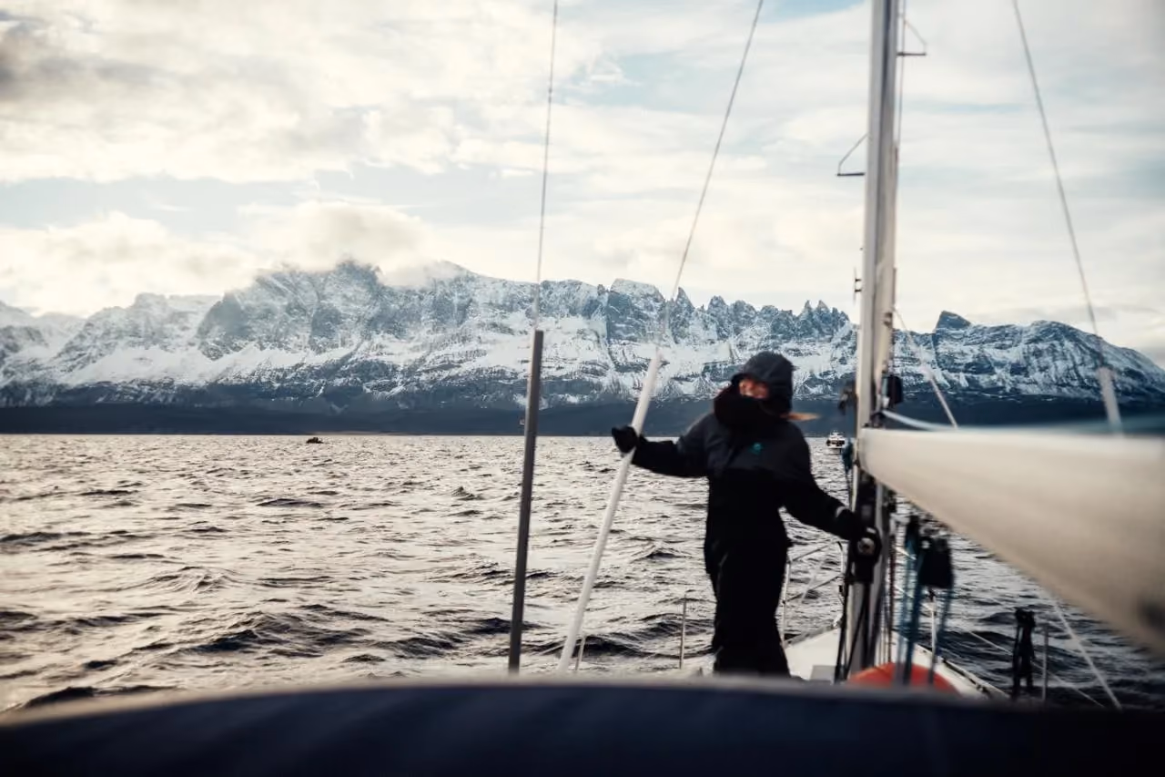 Person standing on a sailboat deck looking out at distant snowy peaks.