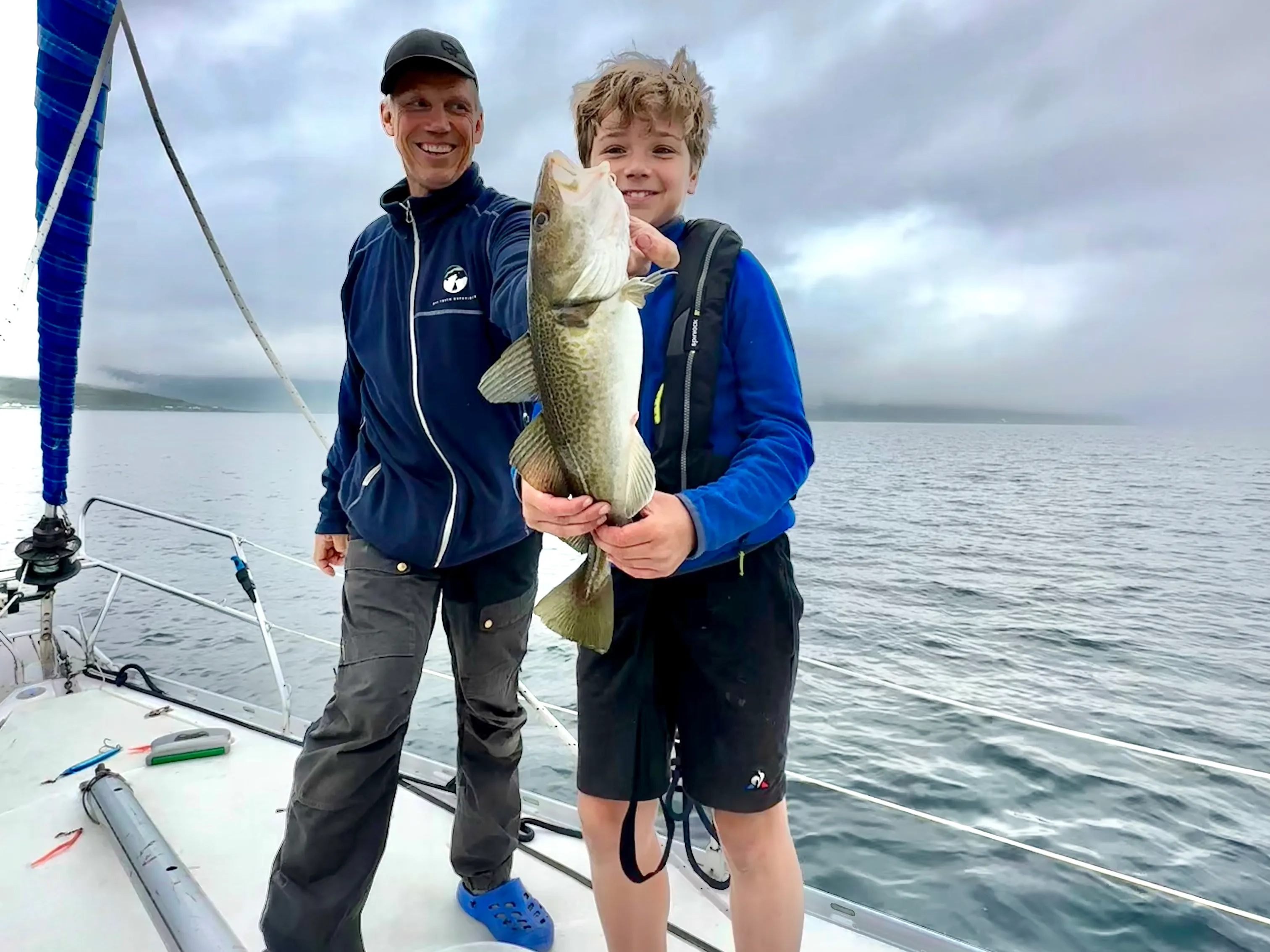 Smiling man and boy holding a large fish on a boat in cloudy waters.