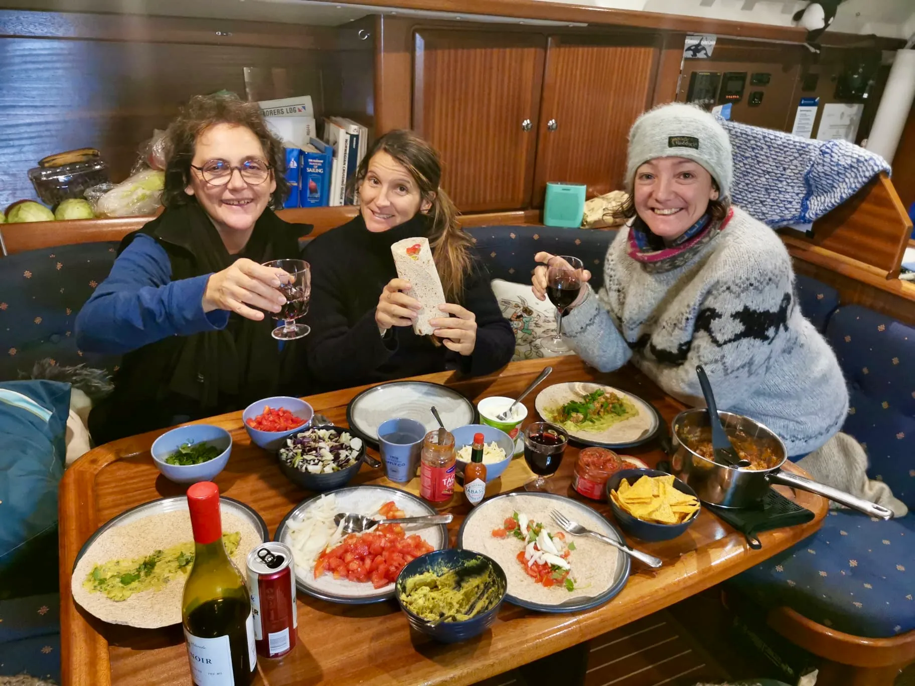 Three women smiling and holding glasses of red wine around a table with Mexican-style food on a boat.
